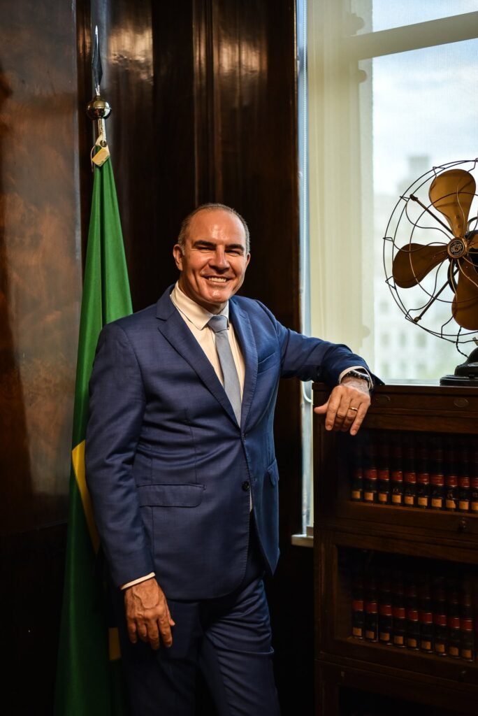 Gustavo Fernandes, advogado, ao lado da bandeira do Brasil, sorrindo