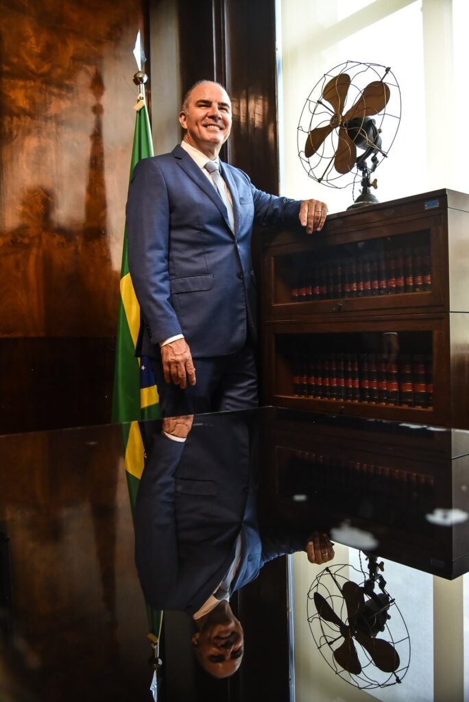 Gustavo Fernandes, advogado full service, ao lado da bandeira do Brasil e estante com livros jurídicos